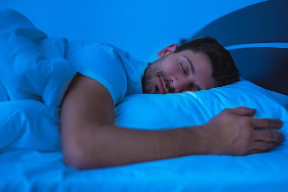 A man sleeps on a bed with white covers; concept of using the Dodow sleep aid to fall asleep.