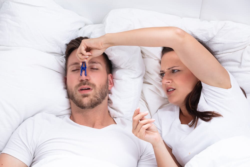 Frustrated Young Woman Trying To Stop Man's Snoring With Clothespin On Bed .