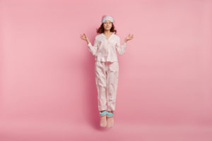 A woman meditating and wearing pink pajamas and standing in front of a pink background.