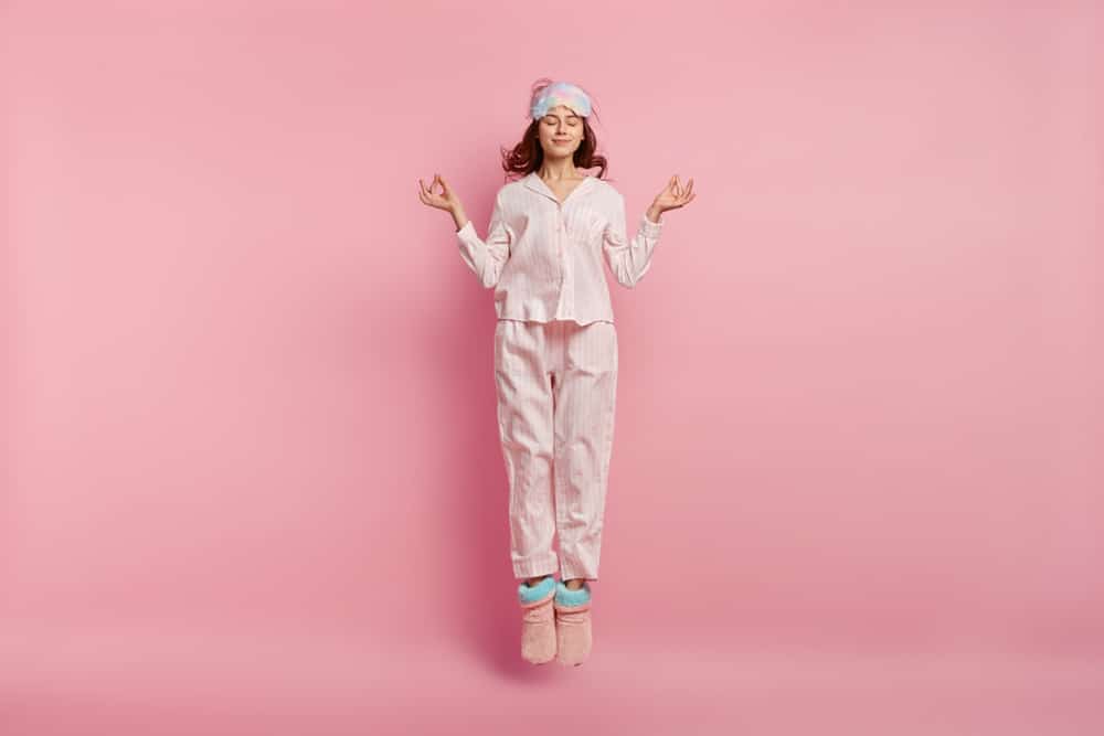 A woman meditating and wearing pink pajamas and standing in front of a pink background.