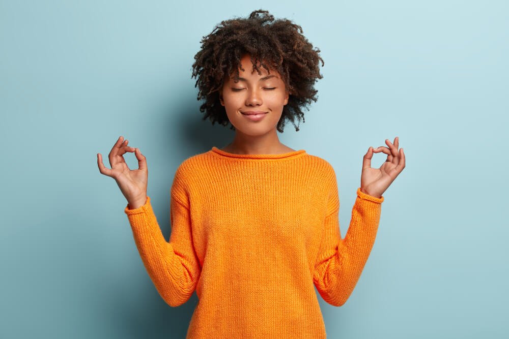 Mindful peaceful Afro American woman meditates indoor, keeps hands in mudra gesture, has eyes closed, tries to relax after long hours of working, holds fingers in yoga sign, isolated on blue wall
