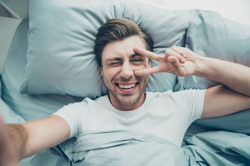 Close up photo of cheerful blonde-haired person lying in bed making v-sign photos in room indoors.