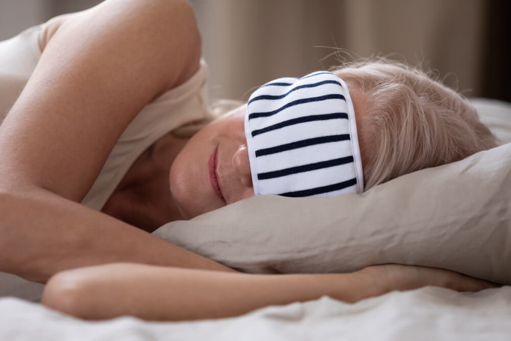 Calm mature woman wearing striped sleeping mask relaxing in comfortable bed close up, peaceful beautiful older female resting in bedroom, enjoying fresh bedclothes, lying on soft pillow