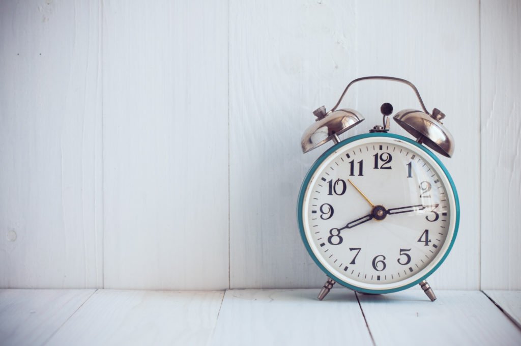A blue alarm clock, with two silver alarms and black hands, sitting against a white wall.