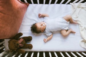 A baby sleeping on a white crib mattress with a teddy bear.
