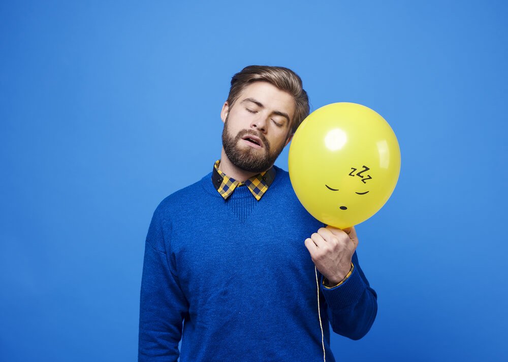 Man sleeping in standing position holding yellow balloon against a blue background.