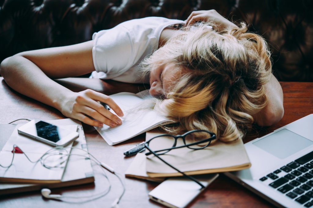 A girl asleep on a desk with a laptop, books, an iPhone and black glasses.