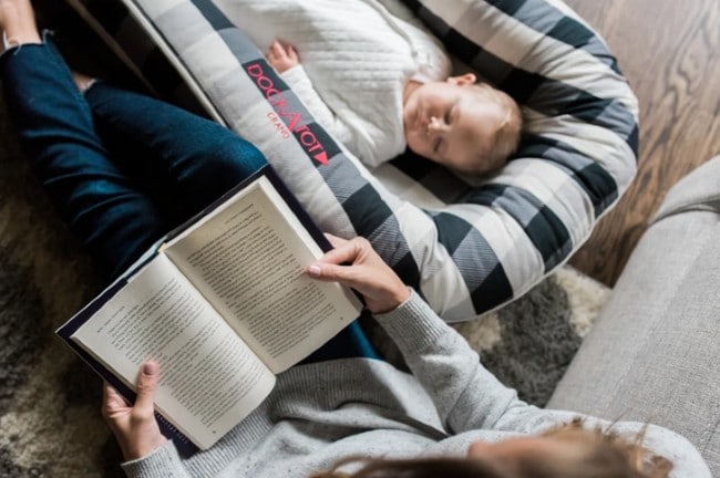 A baby rests while the mother reads a book.