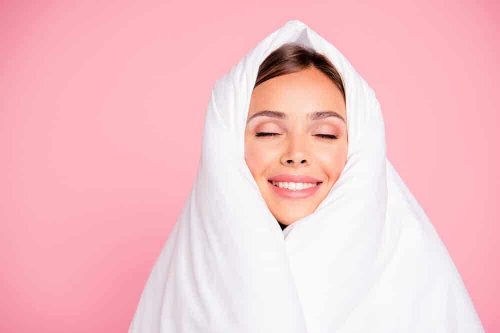 A woman is completely wrapped in a white wool comforter, set against a pastel pink background.