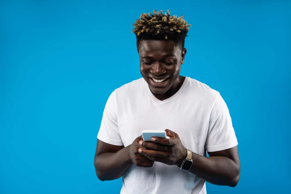 Portrait of young man in white t-shirt using mobile phone and smiling. Isolated on blue background
