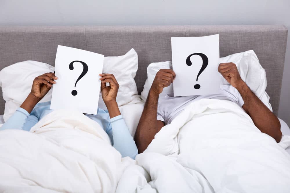 Couple Lying On Bed Holding Question Mark Sign In Front Of Their Face