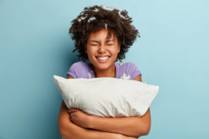 Photo of joyful impressed cheerful woman smiles broadly, has pillow fight, feathers stuck in curly hair, dressed casually, isolated over blue background. Happiness, sleeping and rest concept
