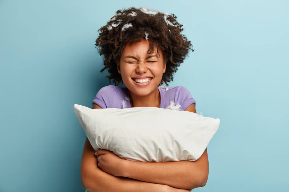 Photo of joyful impressed cheerful woman smiles broadly, has pillow fight, feathers stuck in curly hair, dressed casually, isolated over blue background. Happiness, sleeping and rest concept