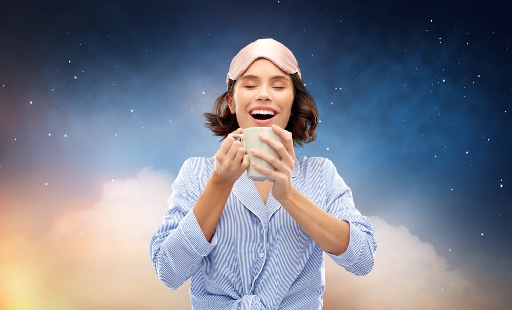 people and bedtime concept - happy young woman in pajama and eye sleeping mask drinking coffee from mug over starry night sky and cloud background; tea for sleep concept.