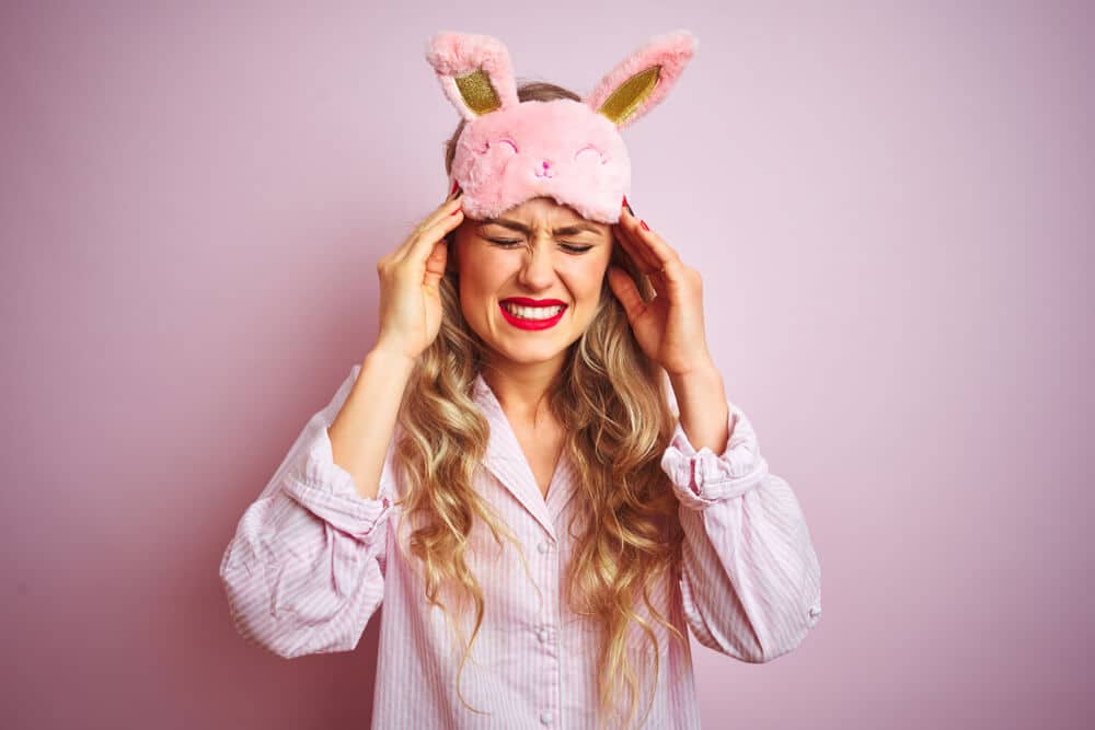 A woman in pajamas holds her hands to her head with a pained expression against a pink background; excessive sleep increases stroke risk concept.