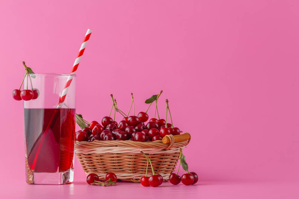 Tart cherry juice in a glass and pitcher on pink with ripe berries in wicker basket