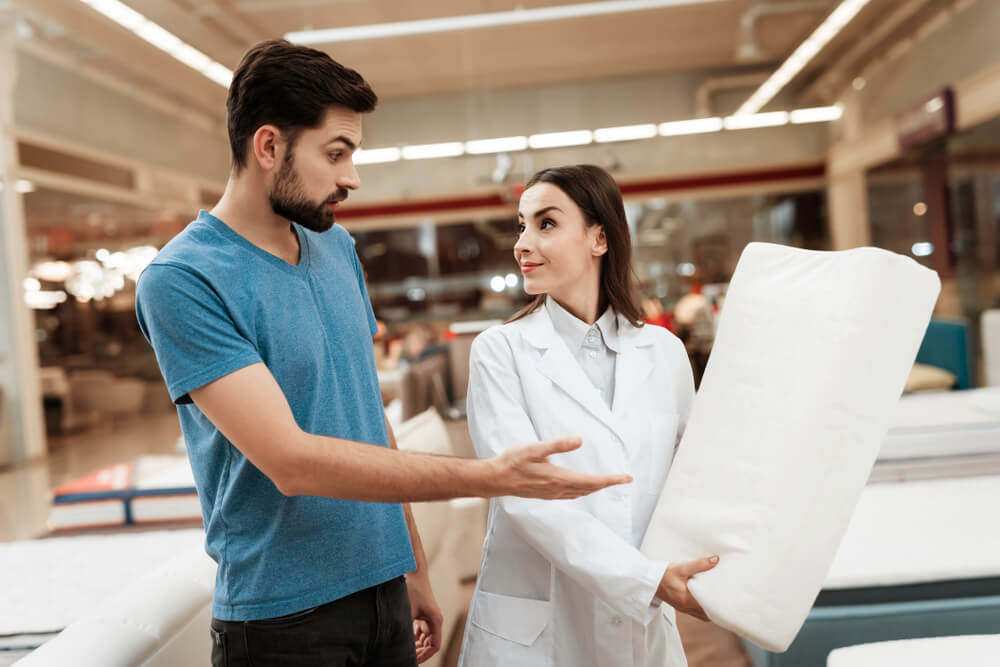 Lovely girl consultant demonstrates orthopedic pillow to confident man in furniture store. Purchase of orthopedic pillow . Choosing pillow in store.