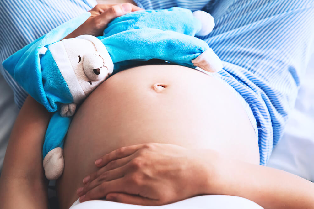 Close-up pregnant woman's belly with teddy toy bear. Beautiful pregnant woman sitting at bed and holds hands on belly in bedroom at home. Pregnancy, parenthood, preparation and expectation concept. N