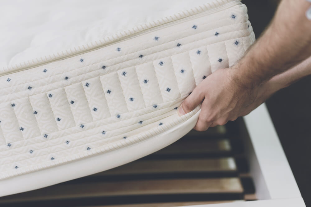 A person setting a white mattress with blue dots on top of a slatted bed frame.