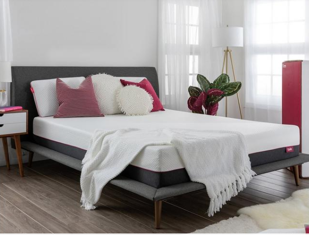 A white Tulo Firm mattress with pink and white pillows, a white throw and a gray mattress frame.