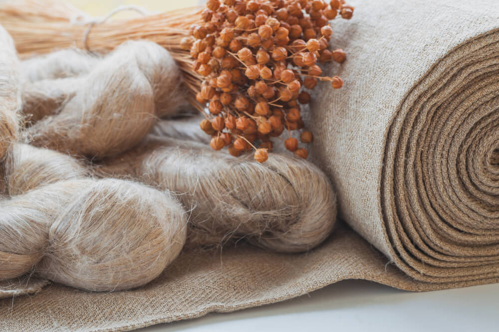 A rolled bolt of flax linen fabric next underneath linen fibers and flax seeds.