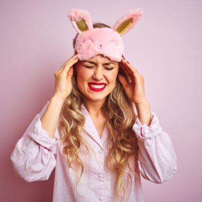 A woman in pajamas holds her hands to her head with a pained expression against a pink background; excessive sleep increases stroke risk concept.
