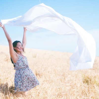 A woman holds a sheet above her head in a field on a sunny day.